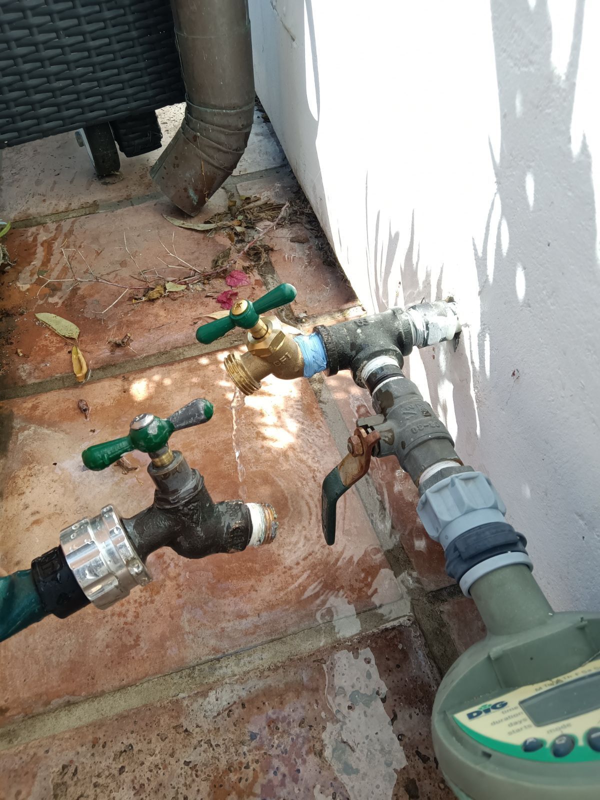 Two outdoor brass faucets attached to a wall pipe system, one connected to a green hose and a digital water meter.