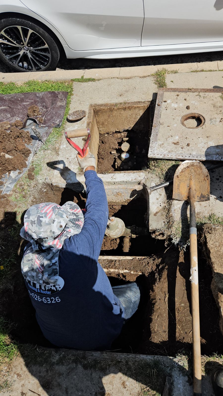 A worker in a patterned hat and blue shirt kneeling in a hole to repair a water meter near a car.