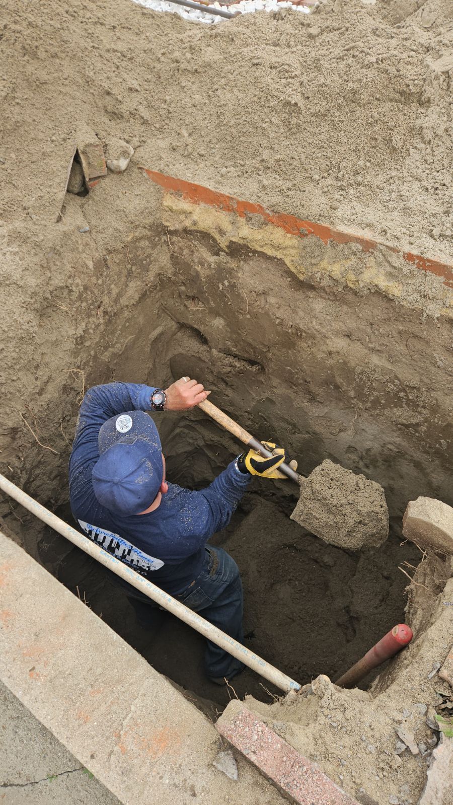 A worker in a blue uniform and cap stands inside a deep, dirt-walled trench, shoveling soil out of the hole.