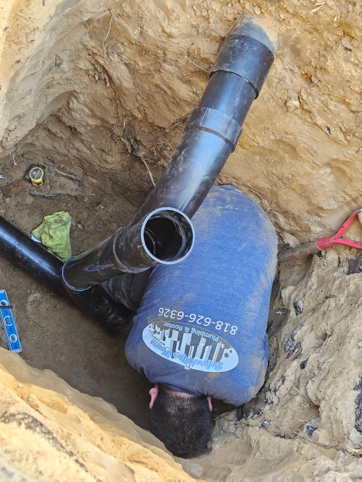 A worker in a blue shirt repairs a black PVC pipe system inside a deep, narrow trench dug into the dirt.