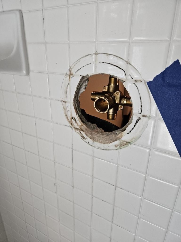 A brass shower mixing valve is recessed inside a circular hole in a white tiled wall.