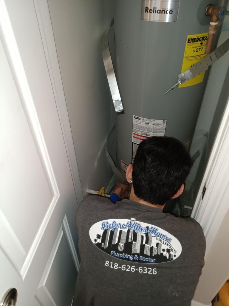 A plumber wearing a gray work shirt repairs a Reliance water heater in a utility closet.