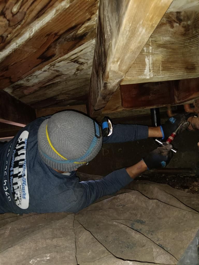A person wearing a beanie, work gloves, and a long-sleeved shirt repairs pipes in a dark, wooden crawlspace.