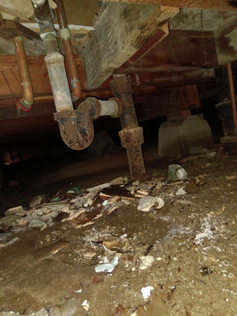 A crawl space showing old, rusted metal plumbing pipes and a P-trap, with debris scattered on the dirt ground below.