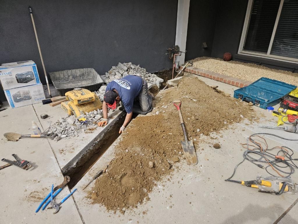 A worker wearing a blue shirt kneels on a concrete patio, digging a trench into the ground beside a pile of dirt and tools.
