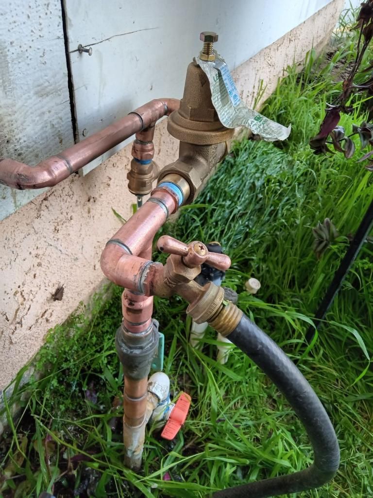 Copper plumbing pipes, a water pressure regulator, and a hose spigot attached to a house exterior above tall grass.