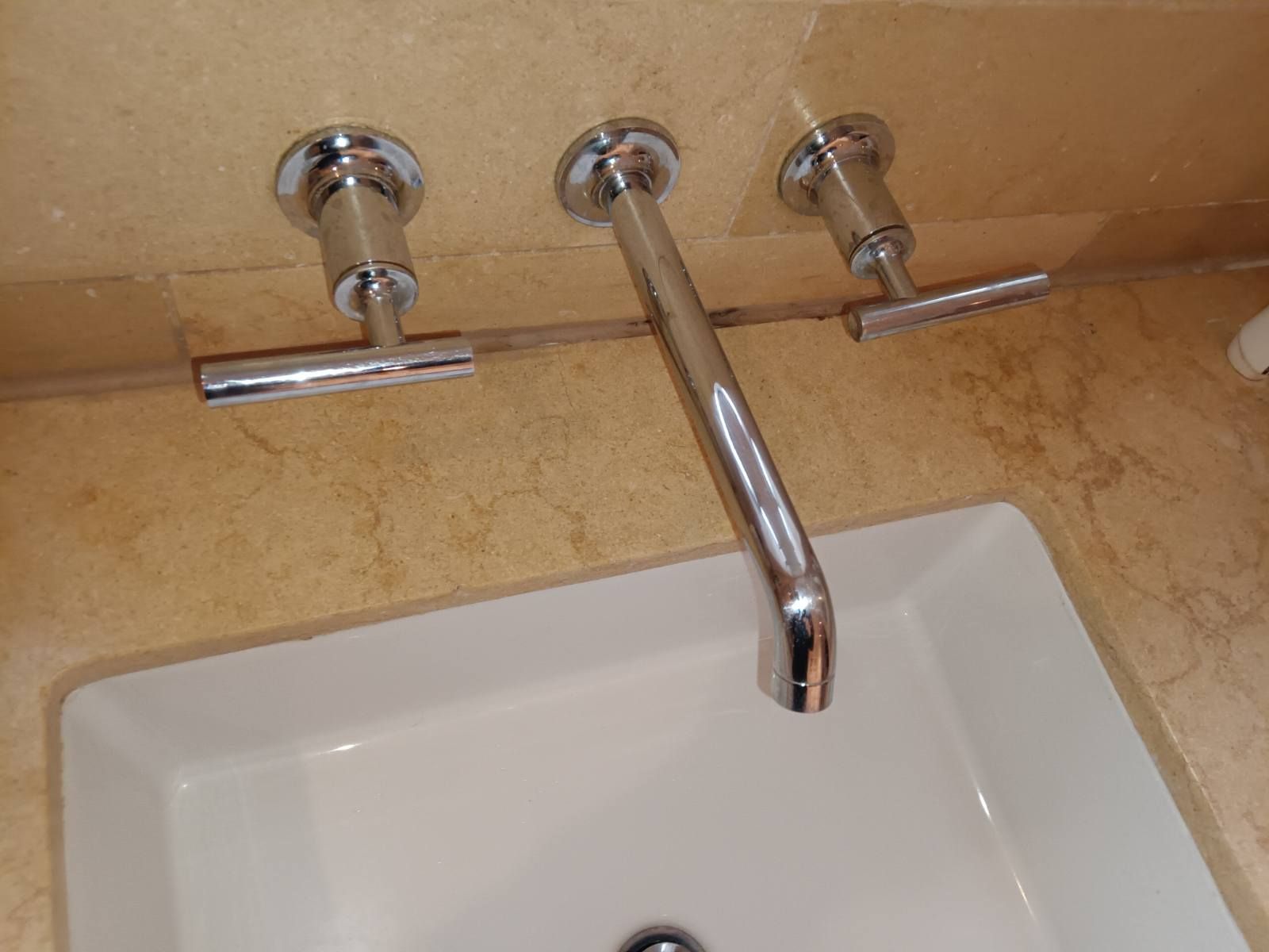 A wall-mounted chrome bathroom faucet with two T-handle controls set against a tan stone backsplash above a white sink.