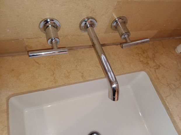 A chrome wall-mounted bathroom faucet with two T-handle controls set against a tan stone backsplash above a white sink.