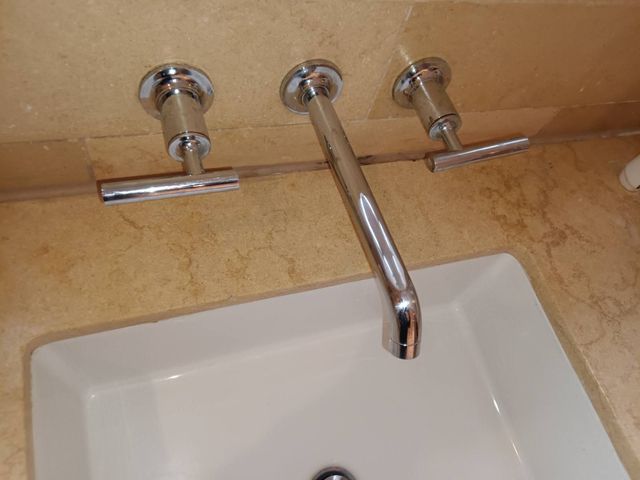 A chrome wall-mounted bathroom faucet with two T-handle controls set against a tan stone backsplash above a white sink.