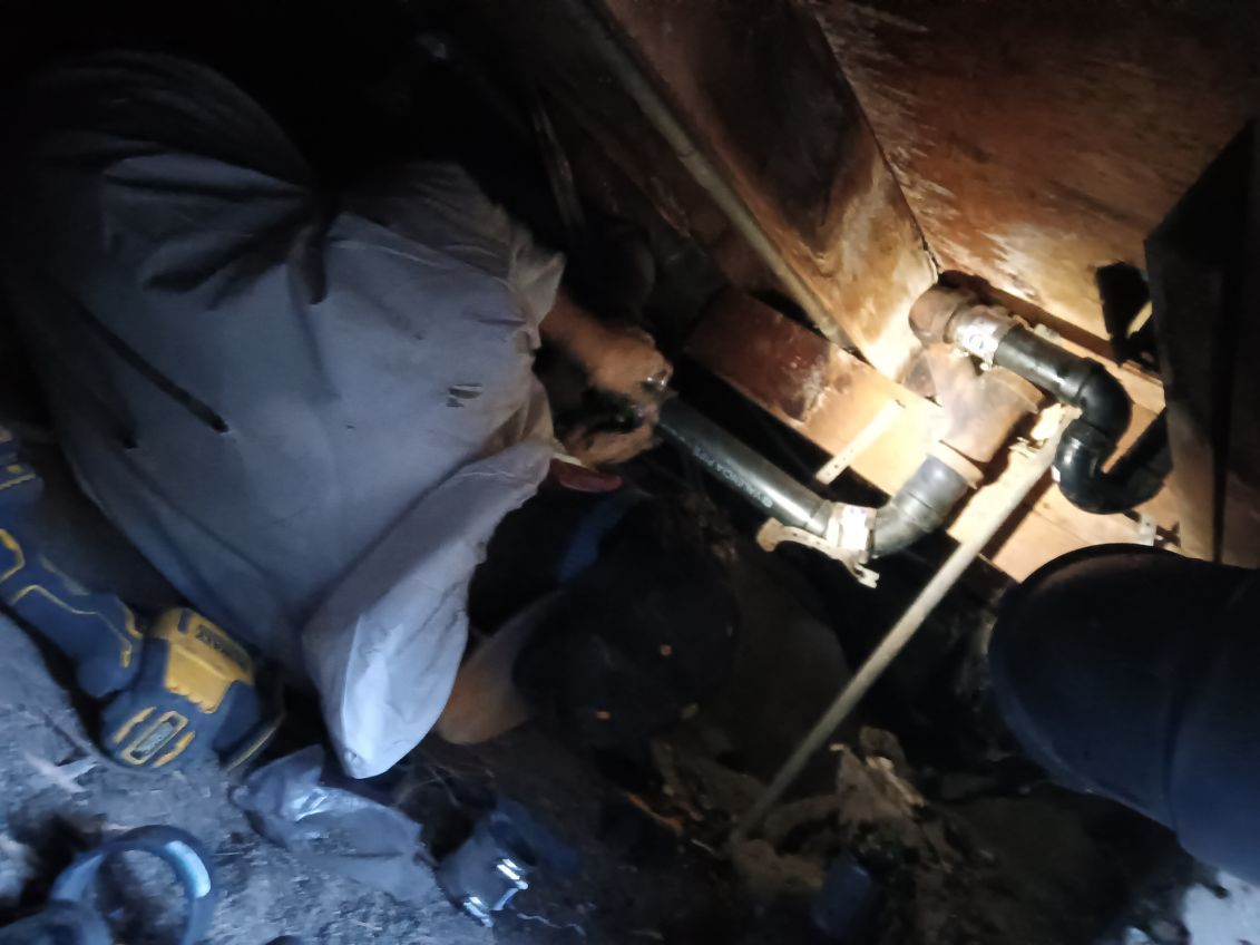 A worker in a dimly lit crawl space repairing plumbing pipes with tools.