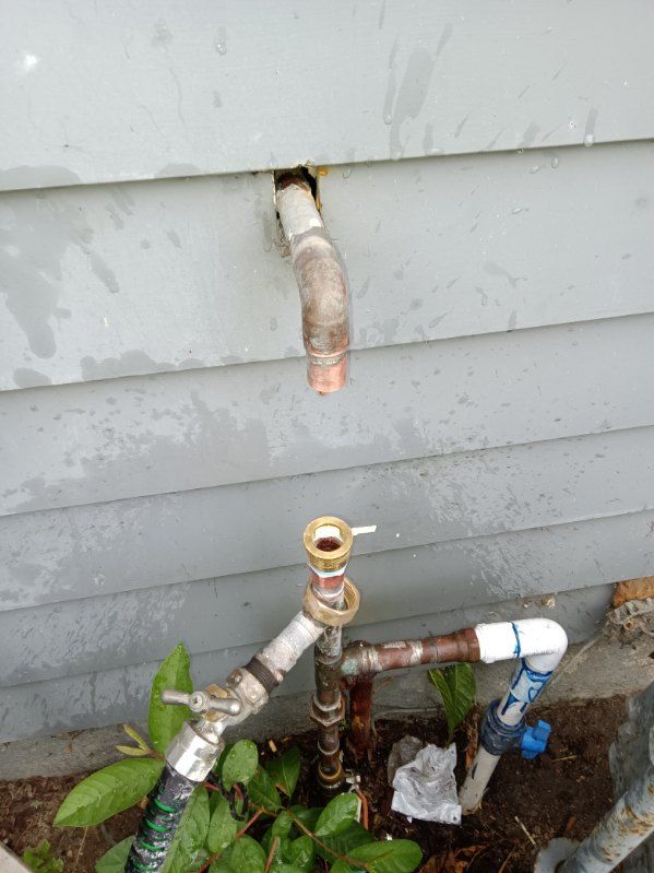 An outdoor wall spigot located above a ground-level assembly of disconnected copper and PVC plumbing pipes.