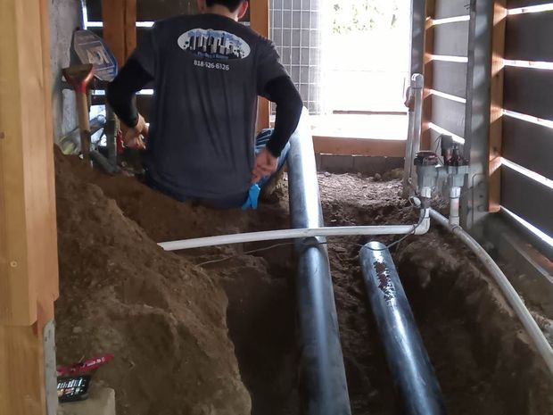A person works in a dirt crawl space, installing black drainage pipes and white PVC conduit near a wooden wall structure.