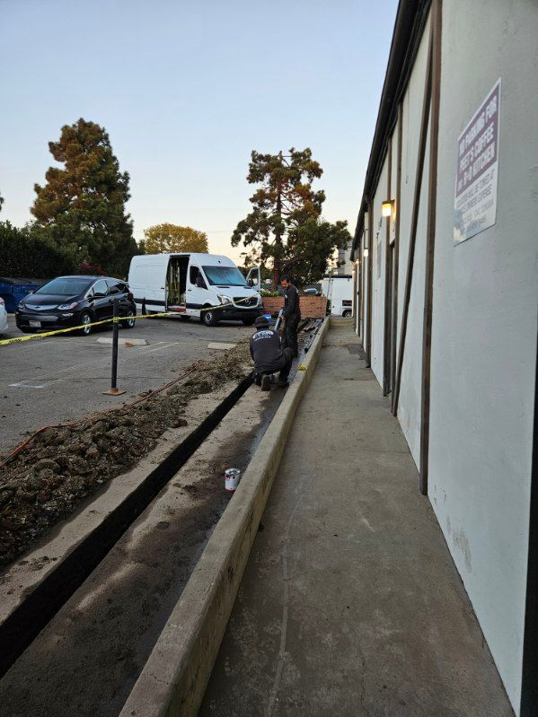 A person in dark clothing moves a wheeled bin along a concrete pathway next to a building trench and parked work vans.