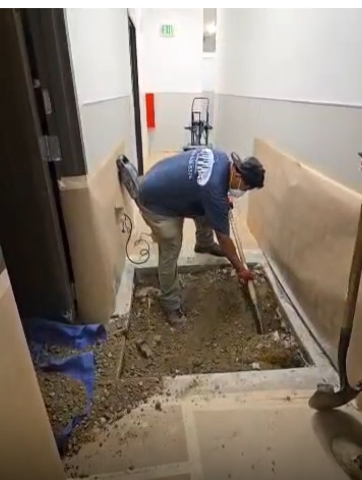 A worker wearing a protective mask digs into a rectangular hole in the floor of an indoor hallway lined with wall paper.