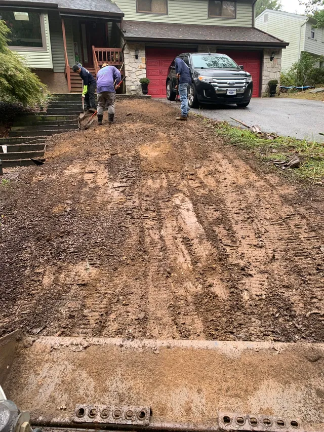 Three workers clear mud and debris from a suburban residential driveway while a dark SUV is parked near the garage.