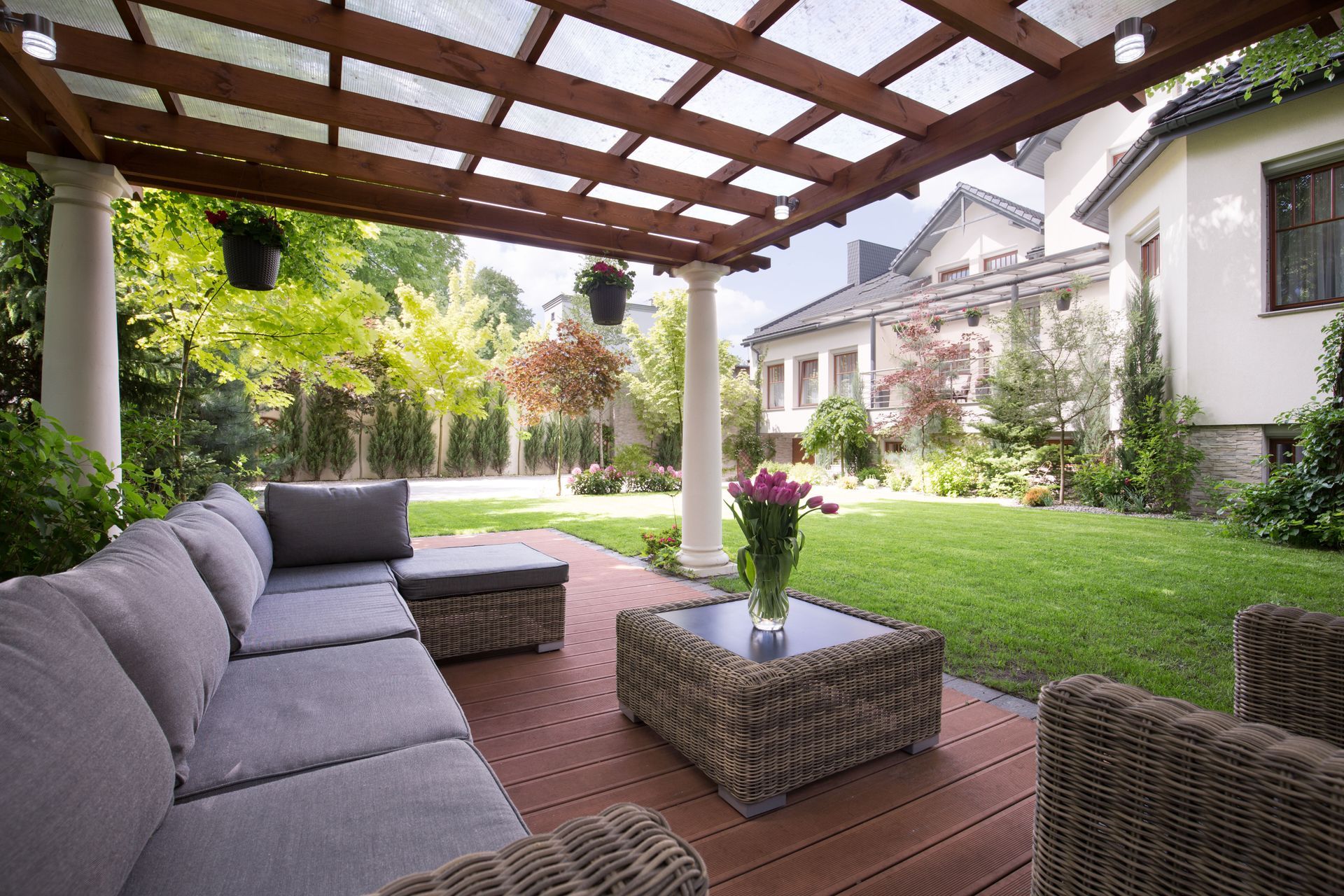 Covered patio with gray sofa, wicker tables, and green lawn beside a white house