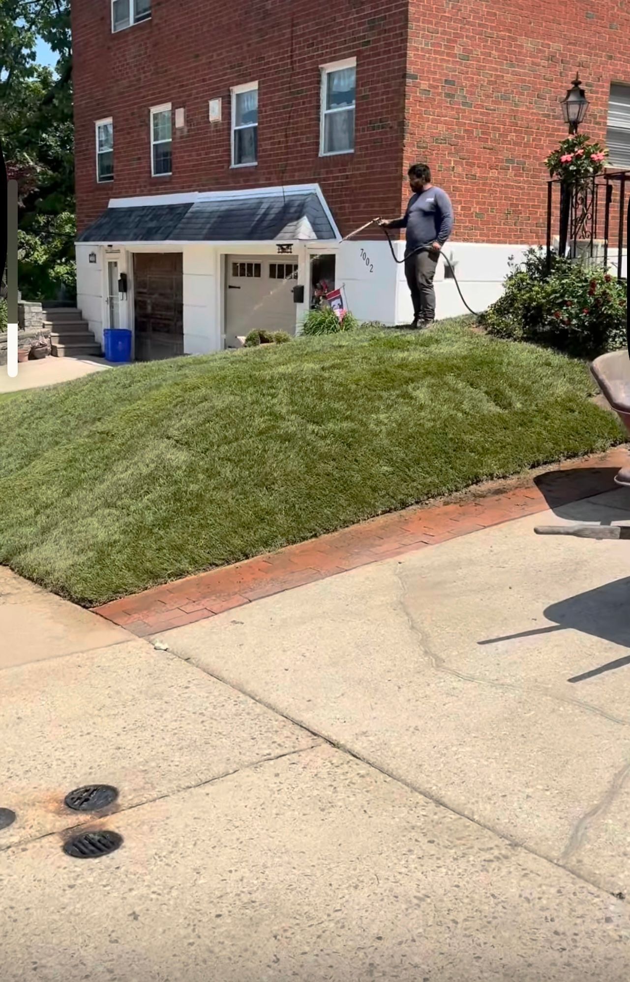A person in dark clothing stands on a grassy slope next to a red brick building, watering the lawn with a hose.