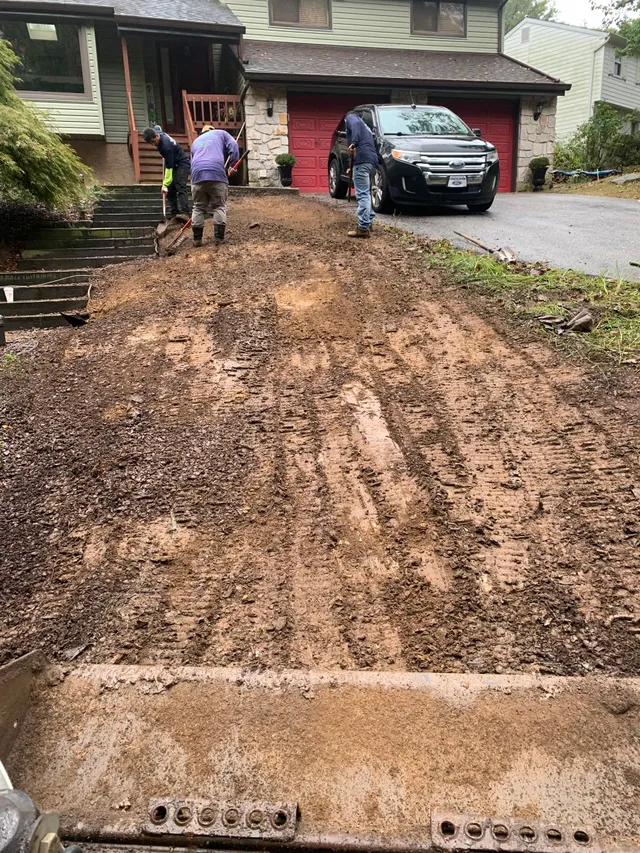 Two workers clearing mud and debris from a suburban driveway toward a house with a parked black SUV.