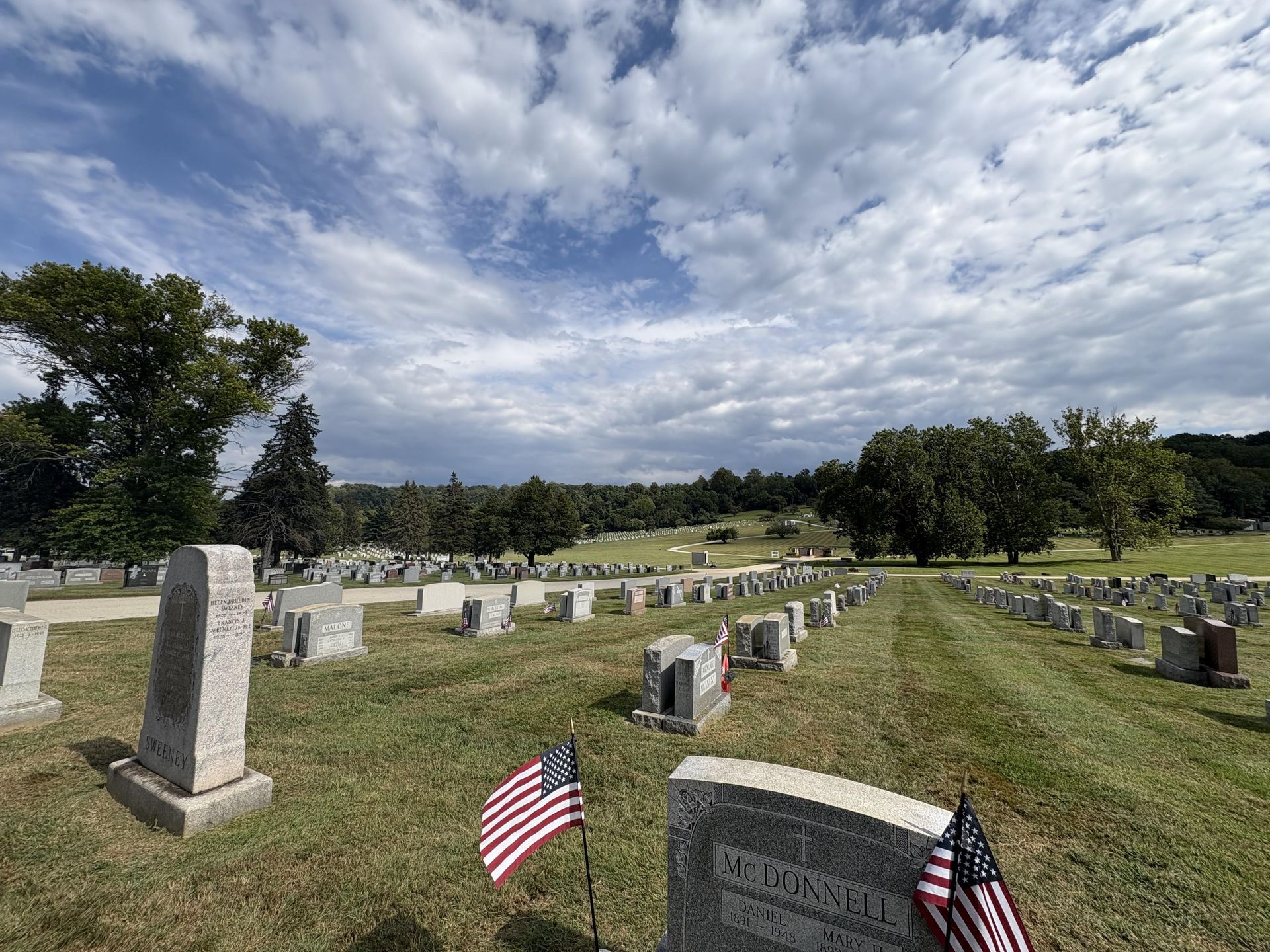 Cemetery with headstones and US flags on a grassy hill under a partly cloudy sky.