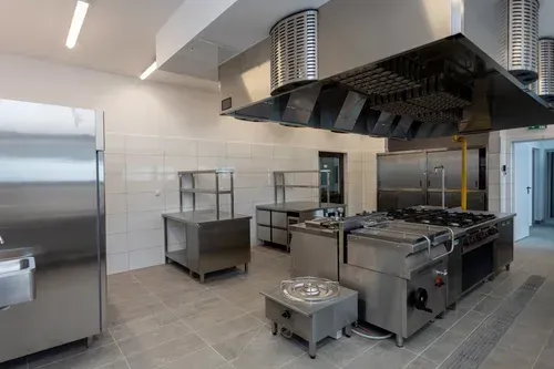Modern stainless-steel commercial kitchen with stove, prep tables, and large ventilation hood