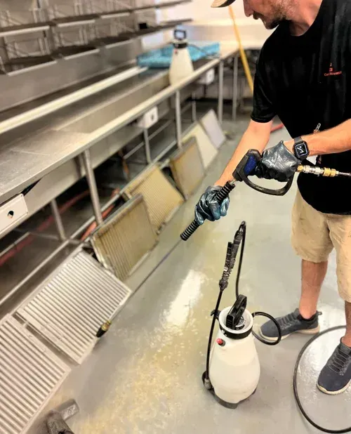 Worker using a spray wand to clean stainless-steel kitchen equipment and floor.