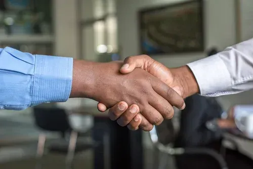 Two people shaking hands in an office setting, one in a blue shirt and one in a white shirt.