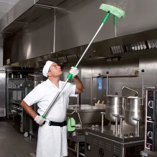 Chef in white uniform cleaning a commercial kitchen ceiling with a long mop