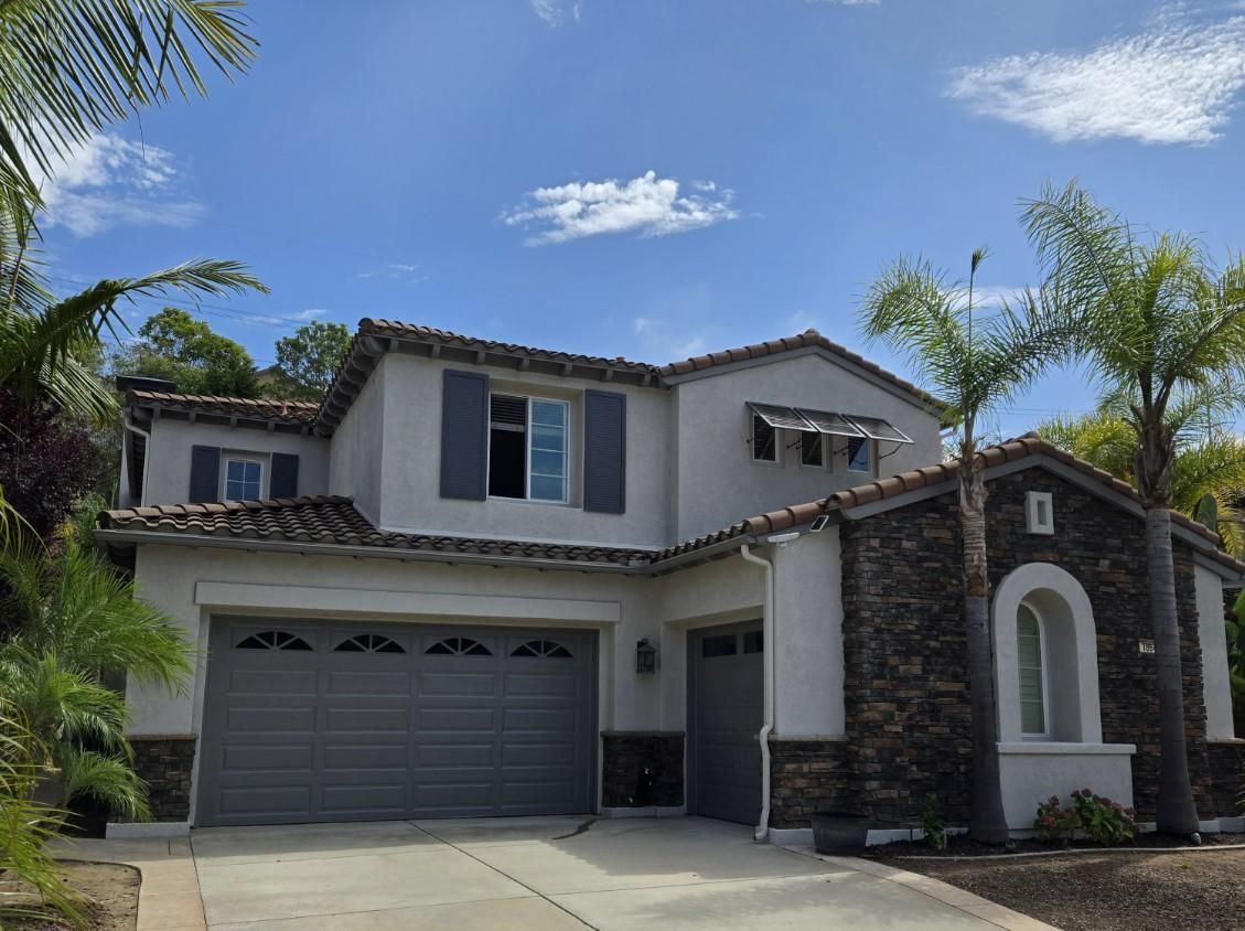 Two-story suburban house with gray garage and stone accents, framed by palm trees under a blue sky