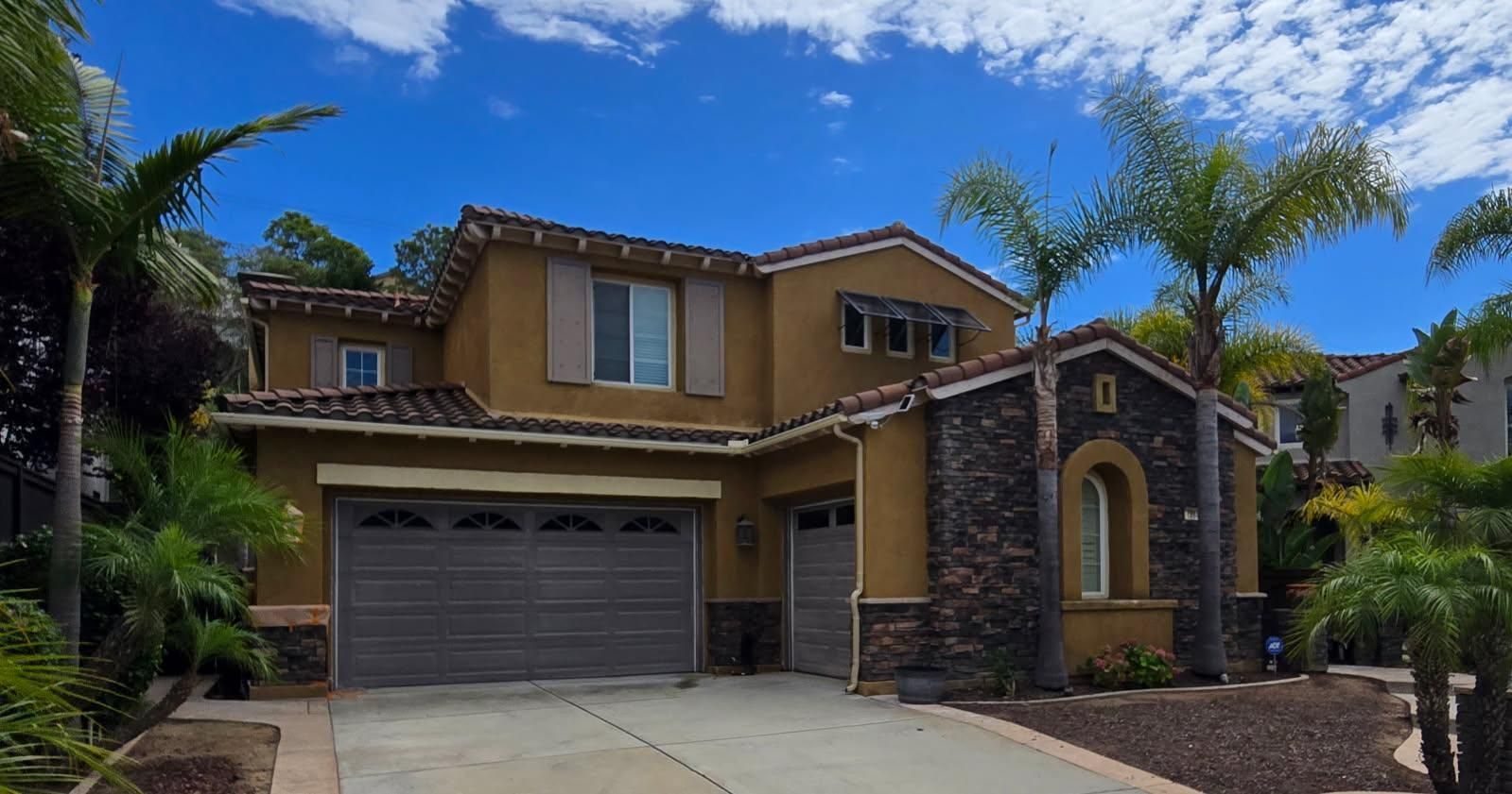 Two-story tan stucco house with stone accents, a dark garage door, and palm trees under a blue sky