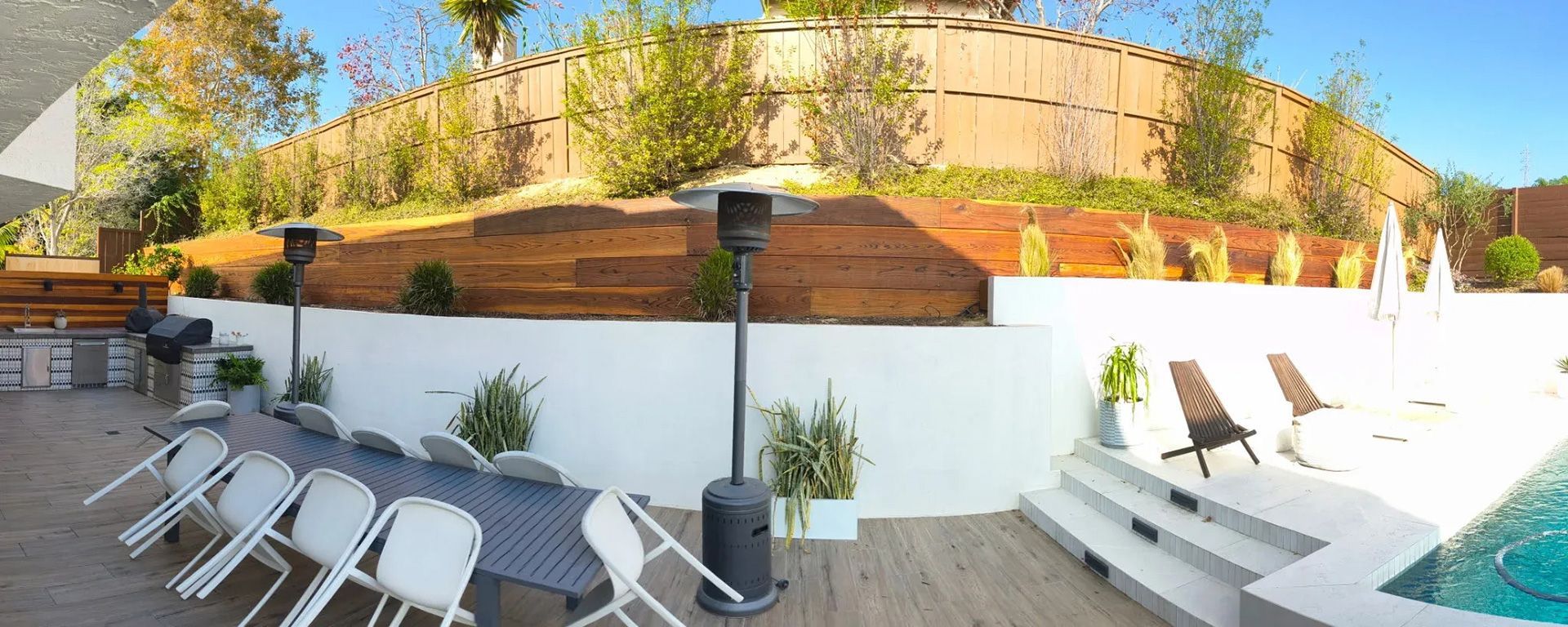 Outdoor patio with white chairs, a long table, a grill, and a swimming pool. A white wall and a wooden fence.