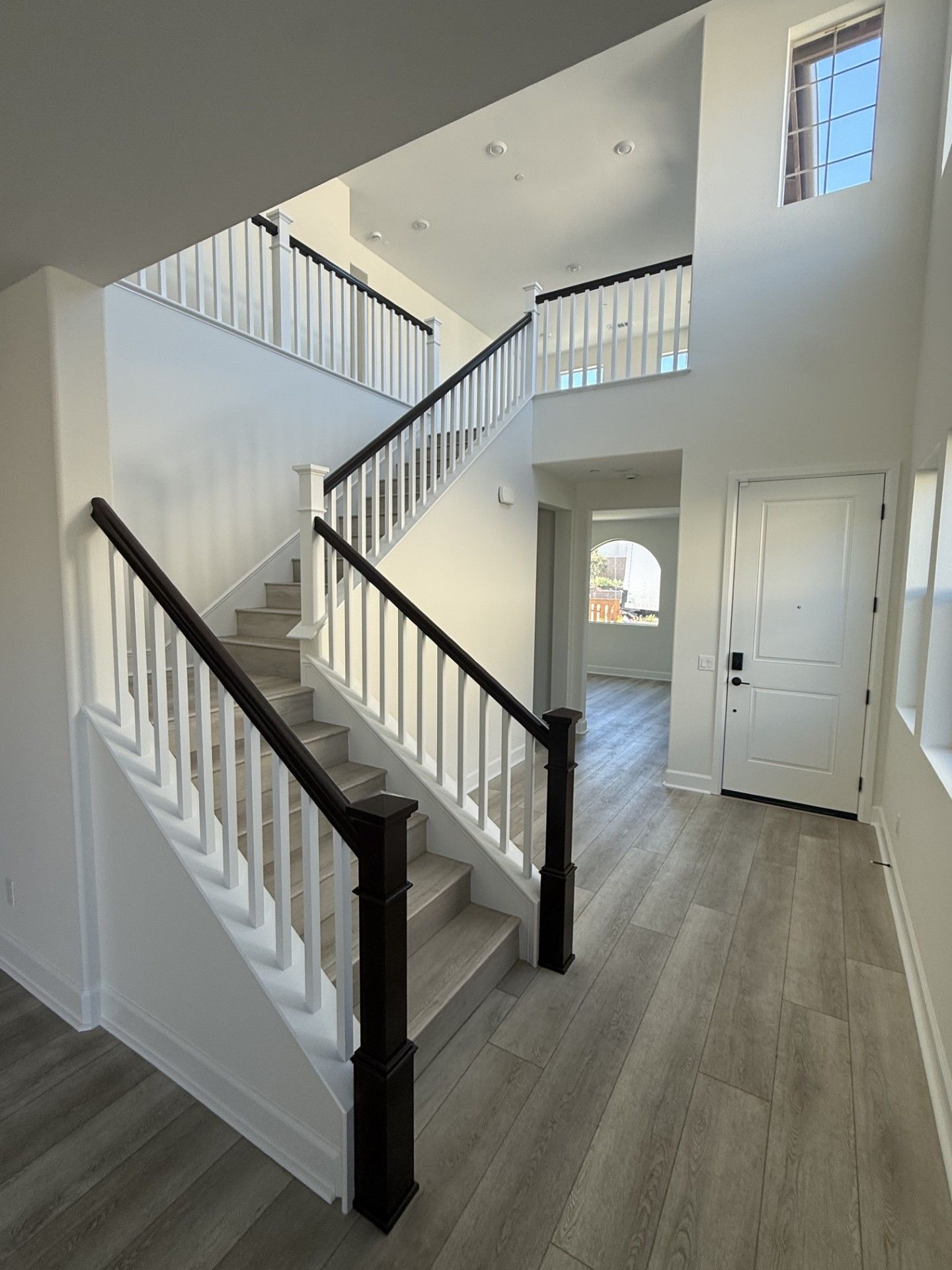 A brightly lit home entryway featuring a staircase with white railings, dark wood handrails, and gray wood-look flooring.