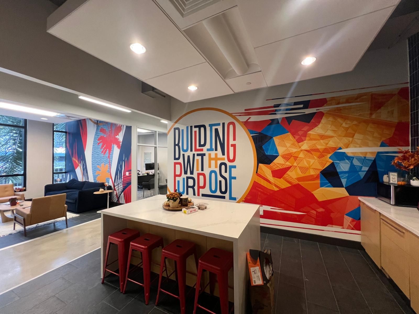 A modern office breakroom with a kitchen island, red stools, and a vibrant mural that reads 