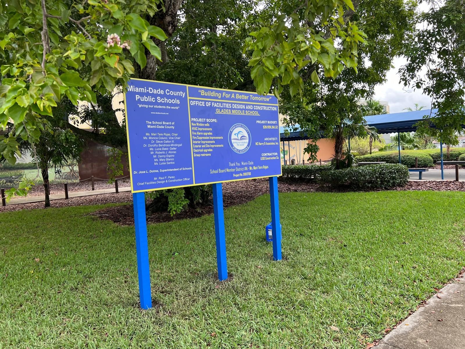 A blue informational sign for a Miami-Dade County park stands on a grassy lawn under a tree.