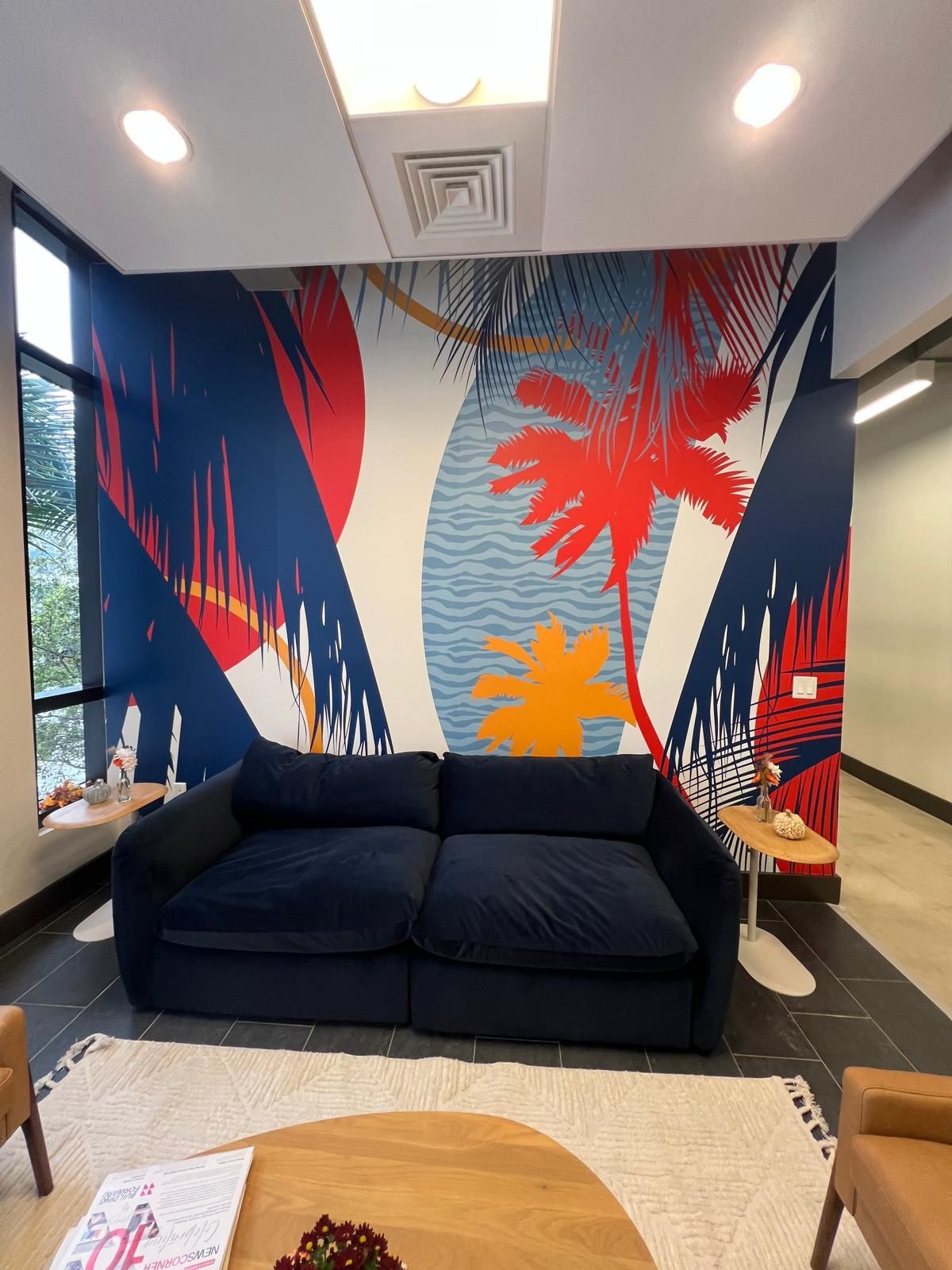 A dark blue sofa sits in front of a colorful, abstract tropical mural on a wall in a brightly lit modern office space.
