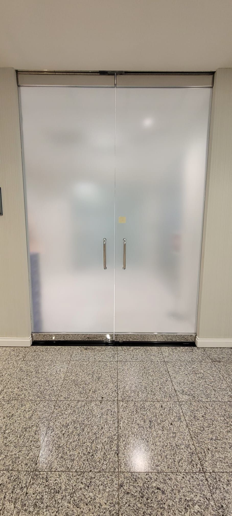 A set of double doors with frosted glass, silver handles, and a metal frame, set against light-colored walls and floor.