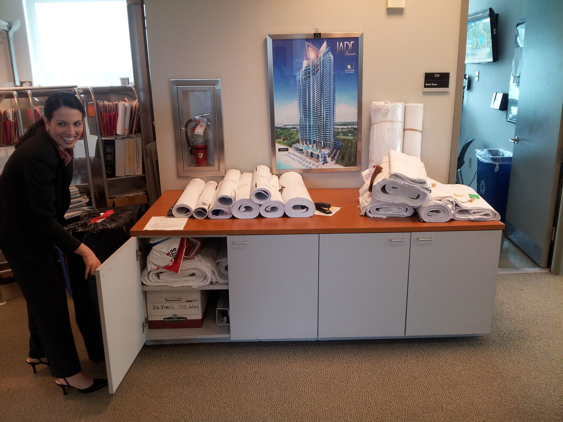 A person in professional attire opens a cabinet door in an office, which is filled with stacked white linens and rolls.