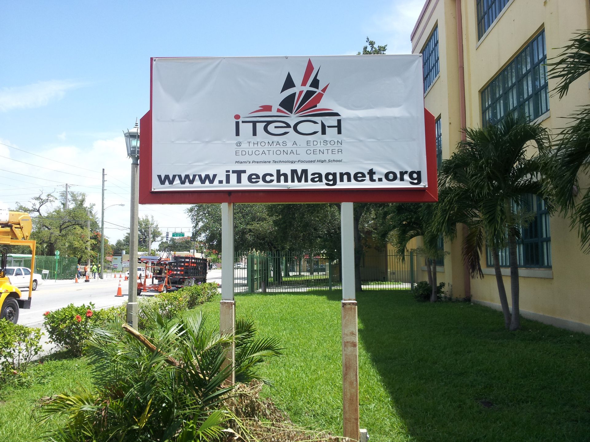 A rectangular sign on two posts displays the ITECH logo and website address in front of a building and trees.