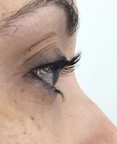 Side-profile close-up of an eye with long eyelashes and a dark eyebrow on a light background