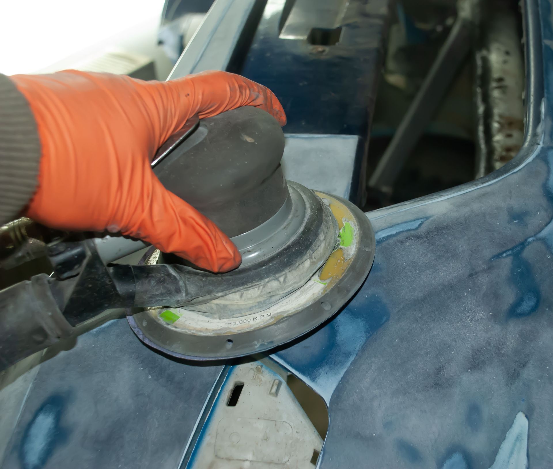 Gloved hand using a sanding tool on a blue car panel in a workshop