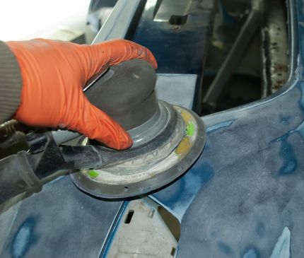 Gloved hand using a sanding tool on a blue car panel in a workshop