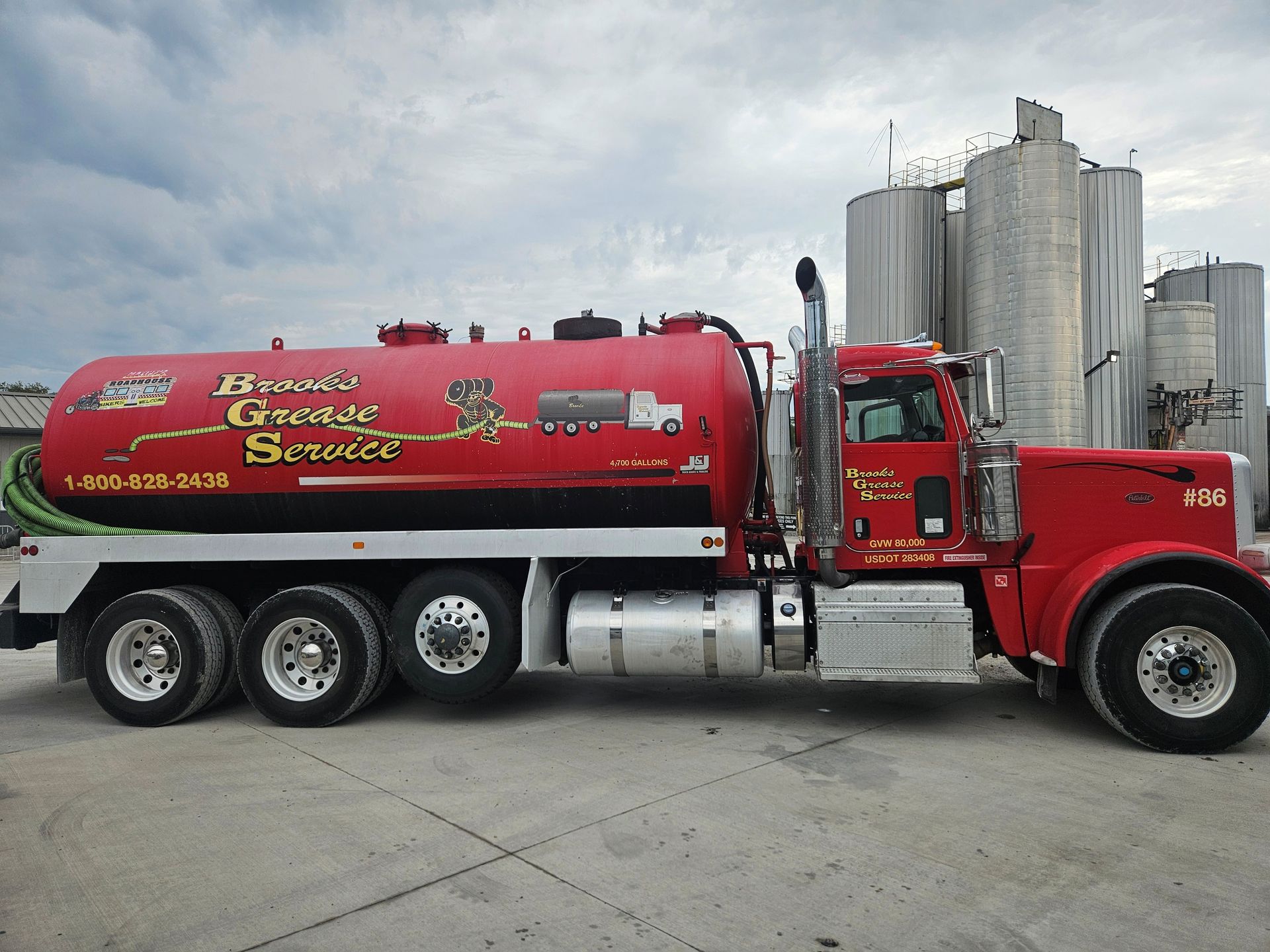 Red septic tank truck with logo, parked near industrial silos, under cloudy sky.