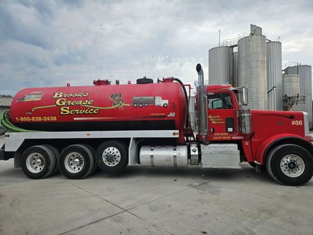 Red septic tank truck with logo, parked near industrial silos, under cloudy sky.