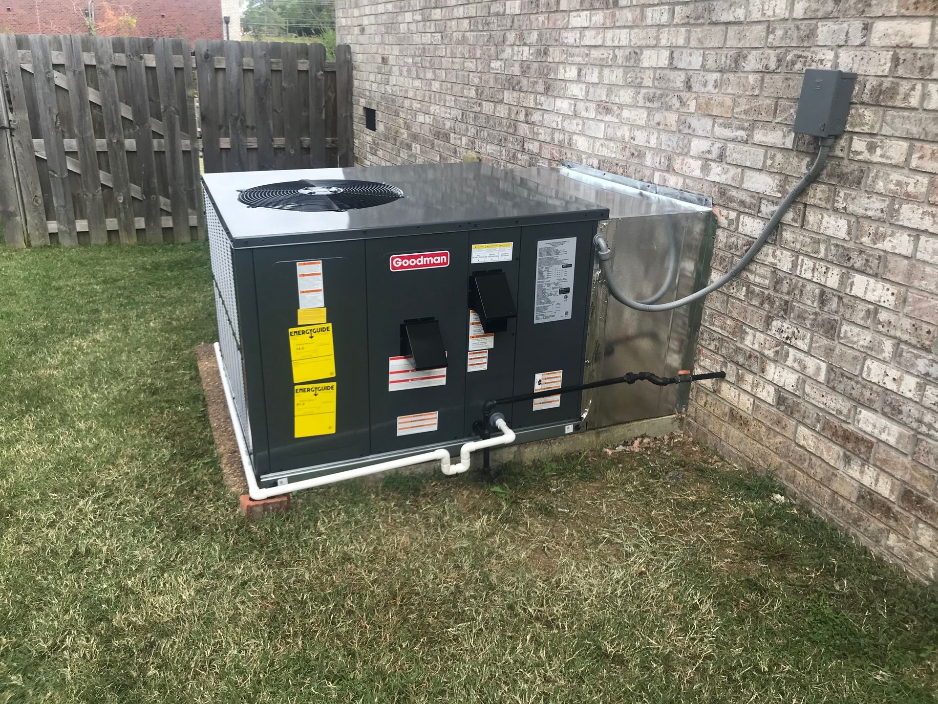 Outdoor HVAC unit beside a brick house wall, with a yellow warning label and fenced yard.