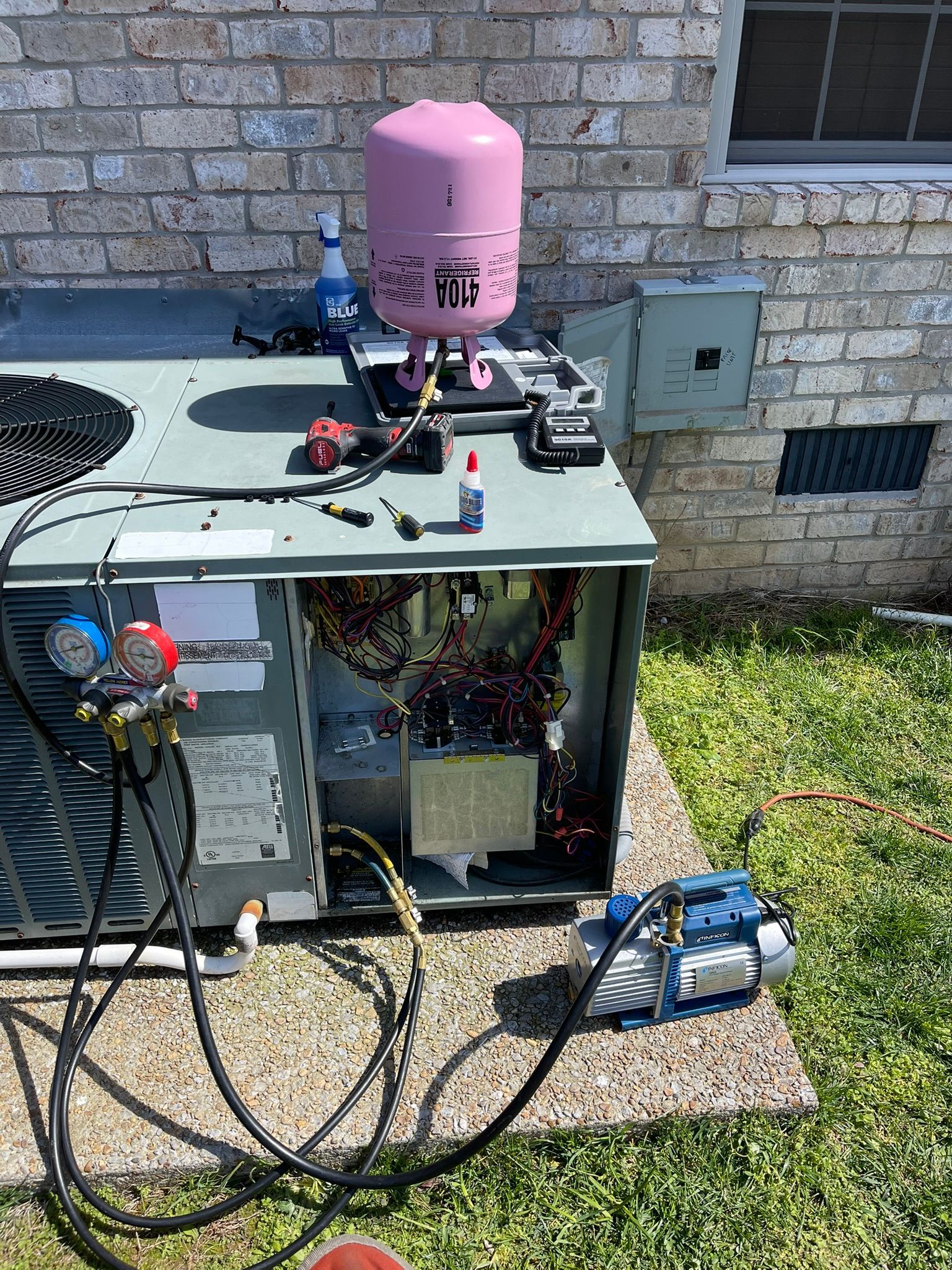 Outdoor HVAC unit with maintenance tools and hoses beside a brick wall and grassy yard