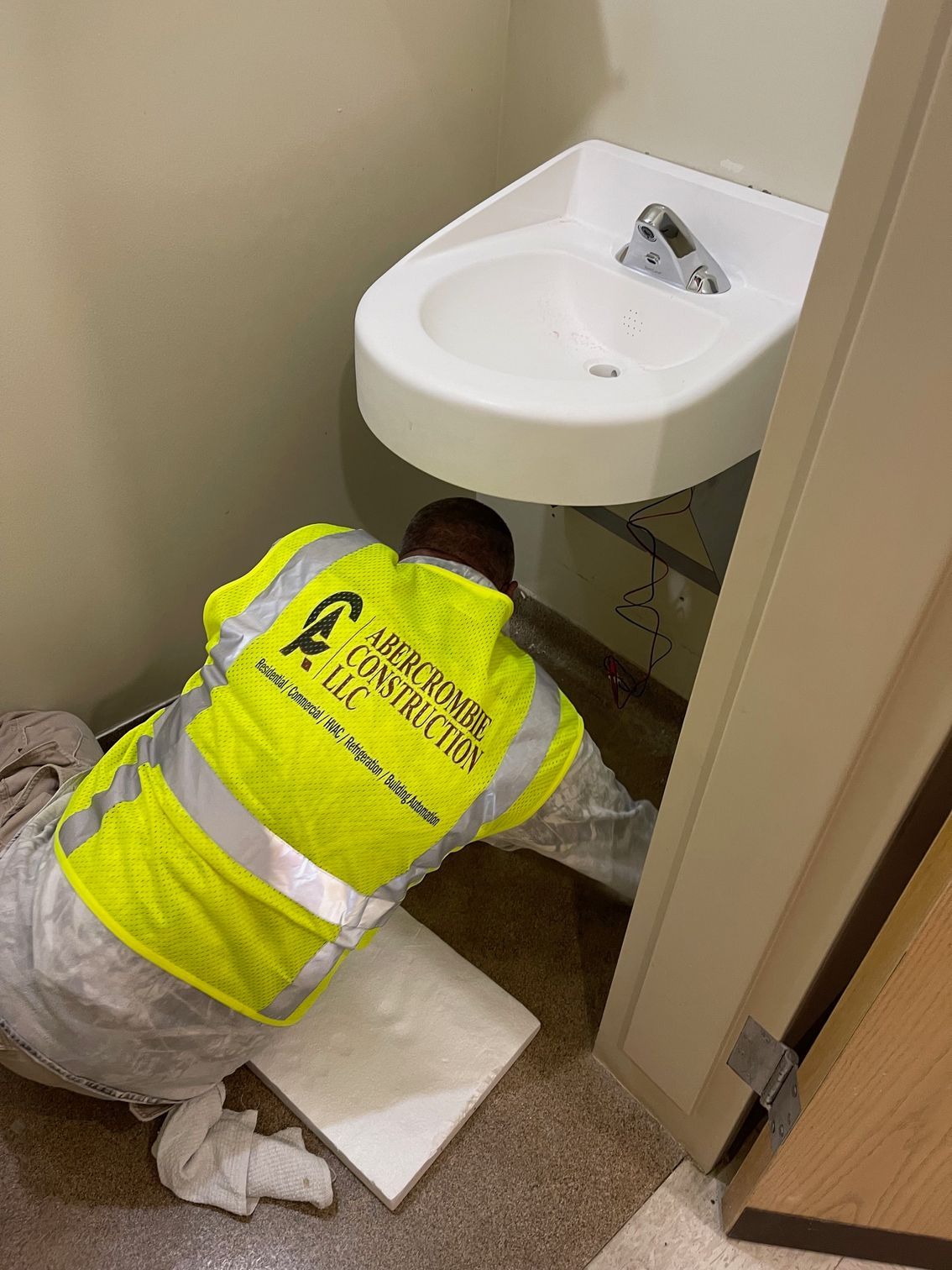 Worker in a yellow safety vest repairing plumbing under a white sink.