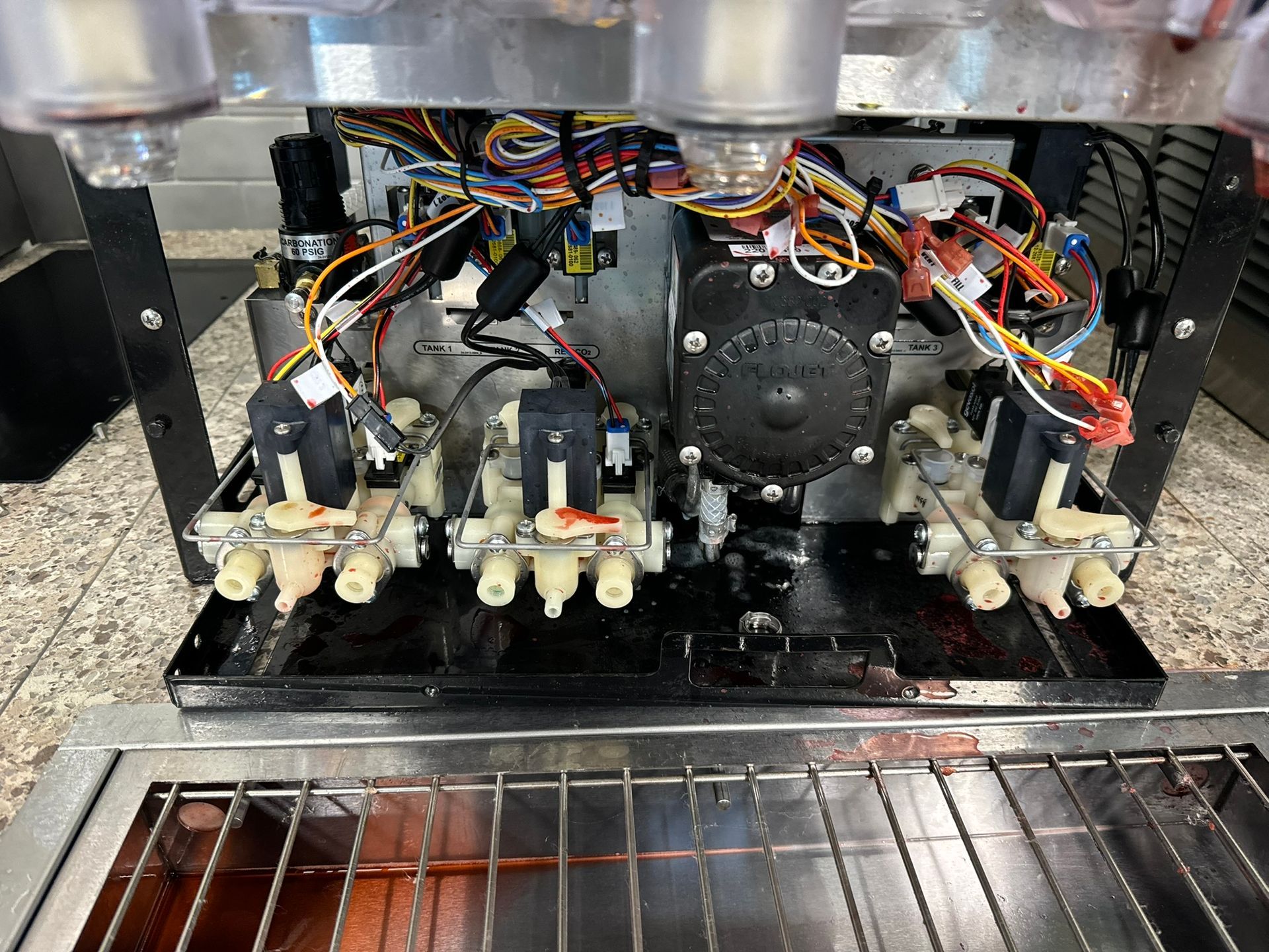 Inside an espresso machine, exposed wiring and pumps above a drip tray and metal grate