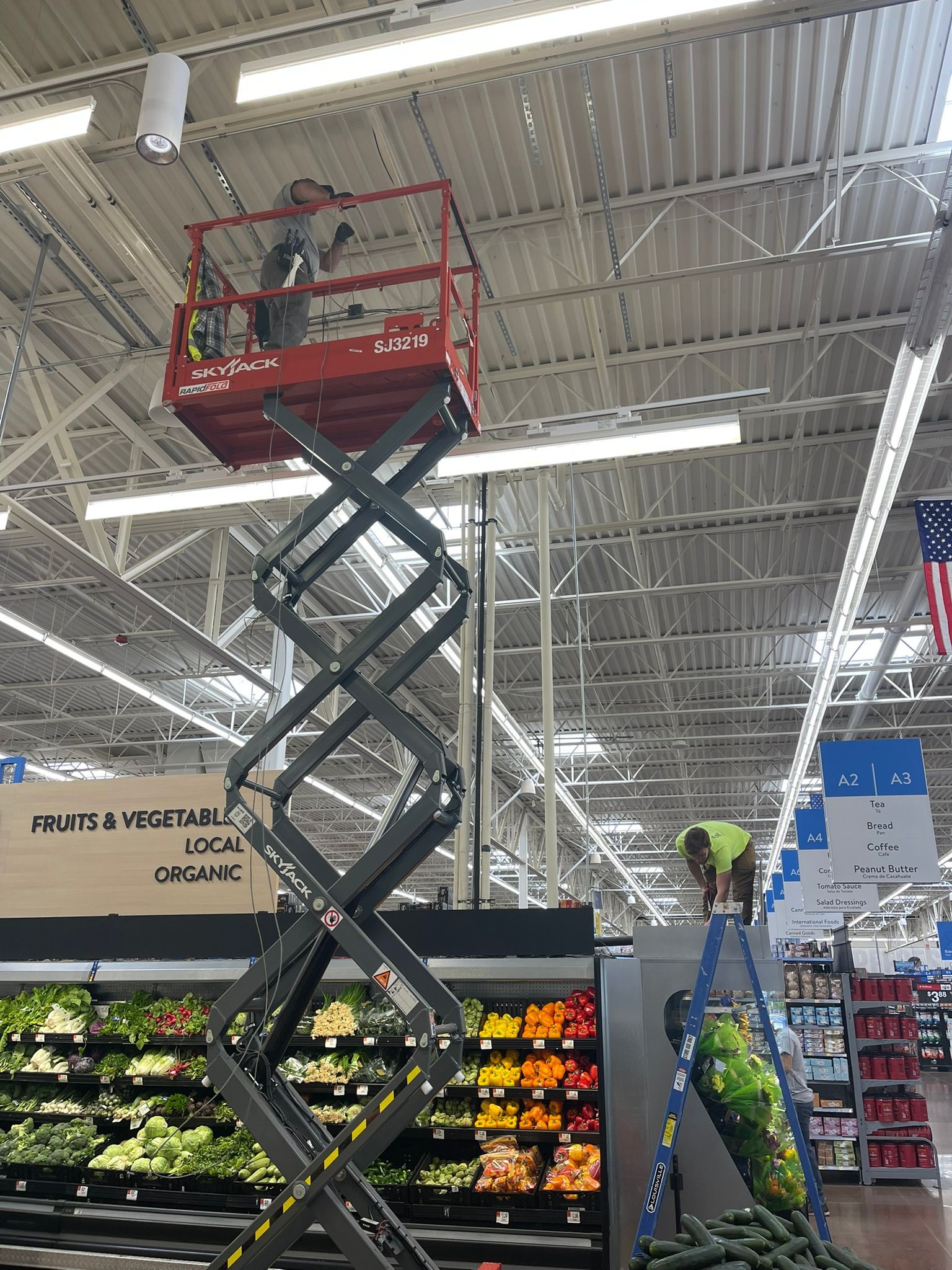 Red scissor lift with a worker in a grocery produce aisle under bright ceiling lights.