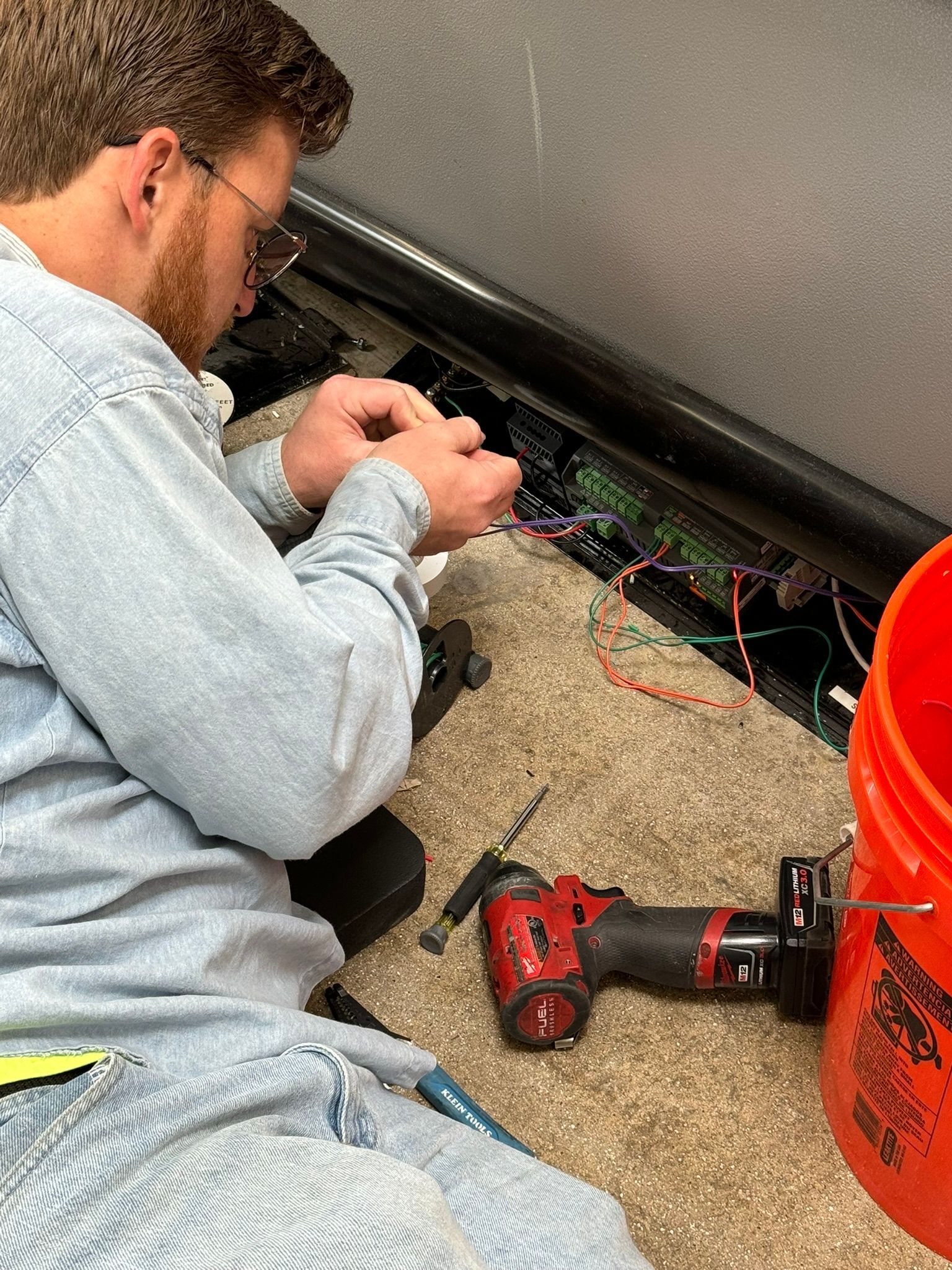 Technician kneels by a wall, wiring cables near a red drill on carpeted floor.