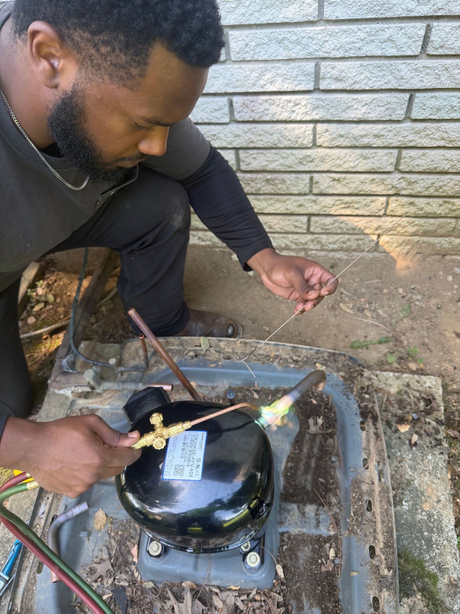 Person repairing a black metal unit outdoors beside a brick wall, using a tool with visible flame.