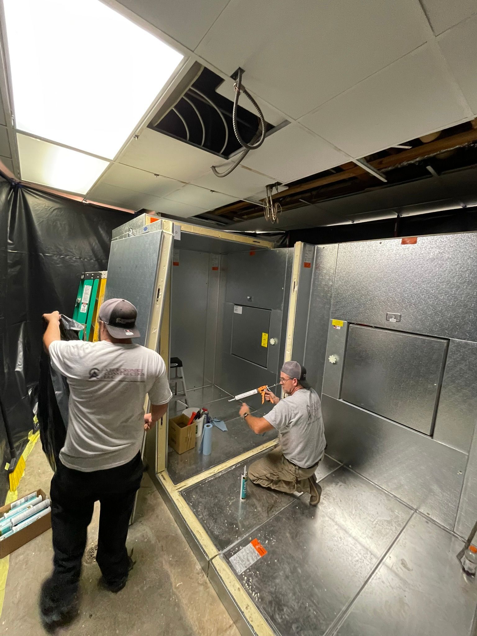 Two workers install silver acoustic panels inside a room with exposed ceiling tiles and ductwork.